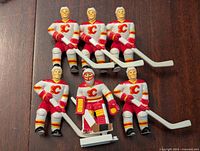 Front view of seven Calgary Flames hockey player figurines including one goaltender, all in white jerseys with red and yellow accents, holding hockey sticks.