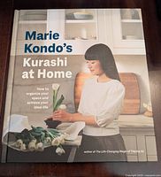 Front cover of Marie Kondo's Kurashi at Home hardcover book showing a woman arranging flowers in a bright kitchen setting.