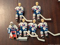 Front view of 6 New York Islanders hockey player figurines in white uniforms, 5 skaters with sticks and 1 goalie with pads and stick