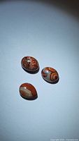 Three oval cabochon cut red crazy lace agate stones on a plain white background, showing distinct swirl and banding patterns.