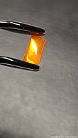 Close-up of the rectangular cut natural Ethiopian fire opal held by tweezers showing bright orange color and inner fire.