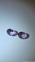 Photo showing two oval-shaped natural amethyst gemstones with slightly different shades of purple, placed on a plain white background.