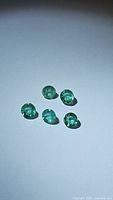 Five natural round emerald gemstones laid out on a plain surface under bright light showing color and size.