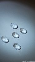Five oval-shaped natural crystal quartz gemstones, fancy rose cut, arranged on white surface with light reflection showing facets.