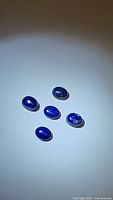 Five oval natural lapis lazuli cabochon stones arranged on white surface with lighting focused on them.