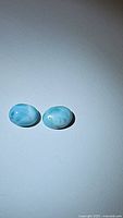 Two oval natural Larimar cabochons placed side by side on a white background showing light blue hues with white marbling.