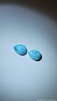 Two natural Larimar cabochons, teardrop shaped, light blue with white marbling, placed on a light background.