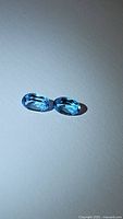 Two oval shaped natural Swiss blue topaz gemstones placed on a white surface showing the blue clarity and facets from different angles.