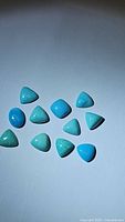 Top view of 11 natural turquoise stones, showing various shapes mostly trillion and some cushion cabochon cuts in light to medium blue color with a smooth polished finish.