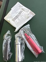 Photo of three aluminum keychain pill containers, red, silver, and black, each sealed in its own clear plastic bag along with a white packaging bag.