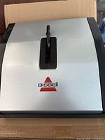 Top view of gray Bissell manual floor sweeper showing body with central handle mount and Bissell logo.