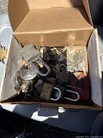 Photo shows open cardboard box filled with 15+ mixed metal locks of various sizes and styles including padlocks with silver, brass, red, and black finishes and multiple keys scattered among them.