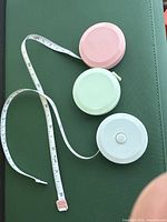Three round retractable tape measures in pink, green, and blue with tape partially extended showing metric and imperial units.