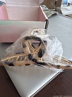 Side view of the beige and black marbled plastic hair clip in plastic wrapping on top of a silver laptop with a box in the background.
