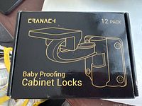 Front of the packaging showing 'Cranach' brand, 12-pack baby proofing cabinet locks with gold text and illustration on black background.
