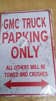 Clear photo of a rectangular metal parking sign with red text on a white background featuring a distressed effect. The text reads 'GMC TRUCK PARKING ONLY ALL OTHERS WILL BE TOWED AND CRUSHED'. The sign has arrows on the bottom and pre-drilled holes for mounting.