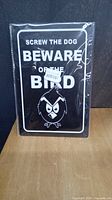Photo of black metal sign with white text 'Screw the Dog Beware of the Bird' and angry cartoon bird illustration, displayed against a dark background on a wooden surface, in original clear plastic packaging.
