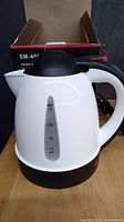 White electric kettle with black handle, lid and water level indicator. Positioned in front of original box.