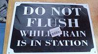 Close-up photo of rectangular metal sign showing text 'DO NOT FLUSH WHILE TRAIN IS IN STATION' in white capital letters on black background, mounted on wooden surface.
