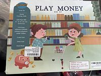 Front image of the Play Money Kids Kit box with colorful cartoon artwork showing children playing and listing the contents on the side: 210 paper bills, 115 coins, credit card, debit card, check book, aged 3+