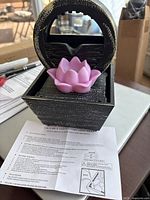 Black battery-operated water fountain with distressed finish, purple lotus flower in front basin, circular water flow area above, displayed on a table with instruction manual below.