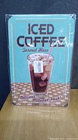Photo shows a rectangular metal sign with an iced coffee glass and retro text on a light blue background.