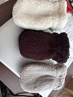 Three fleece infant mittens in light brown, dark brown, and cream laid flat showing front sides.