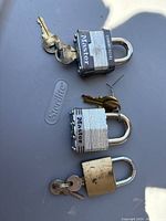 Photo showing three metal padlocks with their keys arranged on a flat surface. The padlocks include a black Master Commercial padlock, a silver Master padlock, and a heavily worn brass-colored padlock.