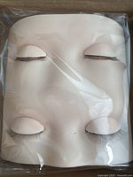 Frontal view of the silicone eyelash practice mannequin head with closed eyes in skin tone, sealed in plastic inside box.