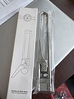 Photo of the multifunction metal bottle opener alongside its original white box with instruction illustrations.