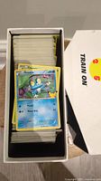 A box filled with many Pokémon cards, showing the top card as a Froakie card with visible stats and artwork.