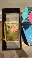 Photo of a large stack of Pokemon cards in a black box, top card is Axew Pokemon card, along with visible black and blue box with Eevee artwork.
