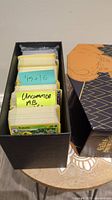 Photo shows black card storage box containing Pokémon cards sorted with handwritten labels, with Kakuna card at the front.