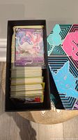 Top view of black box filled with assorted Pokemon cards, Sylveon V card on top and colorful lid with Pokemon illustrations beside.