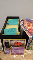 Box containing Pokémon trading cards separated by year labels 13-14 and 15-16 with visible individual cards such as Vulpix and Haunter, and a Pokémon card packaging box adjacent.