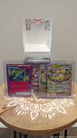 Three Pokemon cards displayed upright in protective sleeves, including Miracle Headset and Jirachi VMAX, on a patterned surface with a white box in the background.
