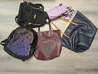 Overview photo of five bags: large black backpack, small geometric black backpack, purple cooler bag, burgundy Roots leather tote, navy blue leather tote.