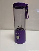 Front view of the purple BlendJet portable blender showing the clear blending container with volume markings, stainless steel blades inside, purple cap with wrist strap, and power button on the base.