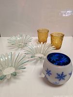 Photo showing 2 amber colored glass candle holders with embossed swirl pattern, 3 blue tinted glass flower-shaped holders, and 1 white frosted glass votive holder with blue snowflakes