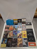 Front view showing 24 cassette tapes arranged with a blue clip-on cassette player on top, displaying various artists' covers.