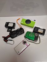 Top view of all cameras and batteries showing Kodak Printomatic green and white, Fujifilm XP green, Pentax Optio black, batteries, and Flip 2000s white and pink