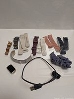 Photo shows Fitbit Charge 6 fitness tracker with 9 interchangeable bands of various styles and a black USB charging cable.