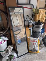 Full-length standing mirror with black frame leaning against wall near various household items including bicycles and bags of salt and soil.