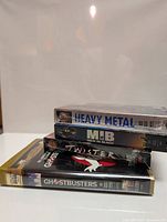 Photo showing four VHS tapes stacked: Ghostbusters, Heavy Metal, Men In Black, and Twister.