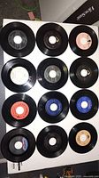 Twelve 45 RPM records displayed showing a variety of labels and colors, several with recognizable brand names and some handwritten markings on centers.