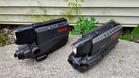 Two vintage black RCA camcorders side by side on a concrete surface outdoors with green plants and siding in the background.