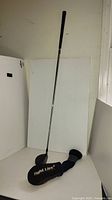 Full view of golf club with black grip and chrome shaft standing upright against white background, black clubhead, and Tight Lies branded sock on the floor.