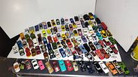 Top-down view of about 80 toy cars arranged in rows on a white surface showing variety of colors and types including sports cars, trucks, and emergency vehicles.