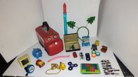 Photo showing an assortment of kids toys including a red bus, blue piggy bank, small toy cars, a fidget spinner, a mini puzzle, and various small toys and figurines laid out on a white surface.