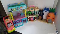View of all toys in the lot arranged on a white surface, including the Moj Moj claw machine, SpongeBob pineapple house, blue underwater tower, green house playset, and orange toy bus.